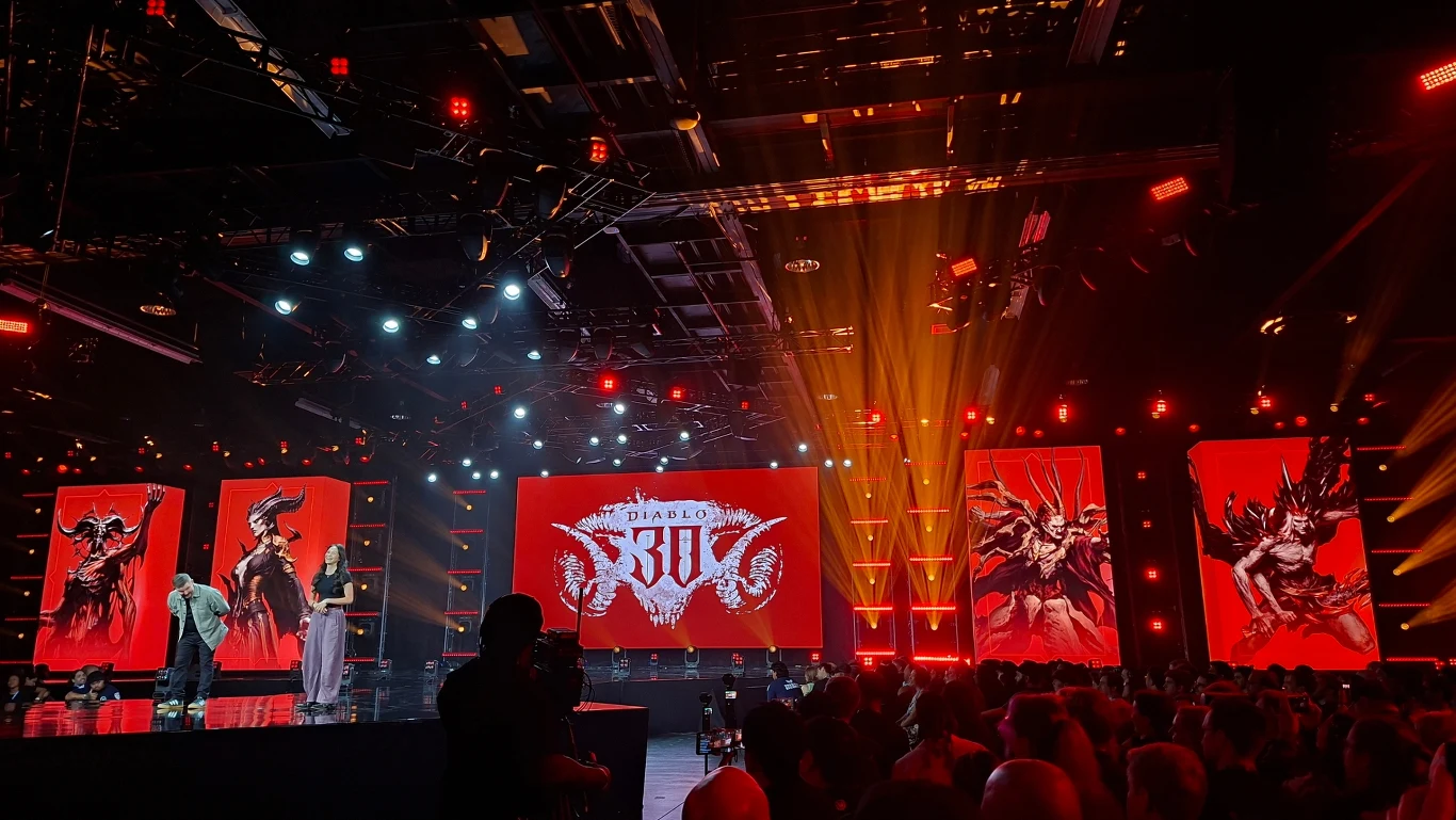 Scena oświetlona czerwonymi światłami, na której dwie osoby rozmawiają na tle trzech ekranów wyświetlających grafiki związane z grą Diablo oraz logo Diablo 3:00, wokół liczna widownia.