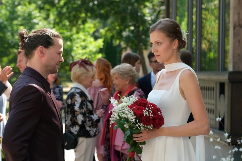 Para młoda stoi naprzeciwko siebie; kobieta w białej sukni trzyma bukiet czerwonych róż, a w tle grupka elegancko ubranych osób rozmawia ze sobą podczas uroczystości na świeżym powietrzu.