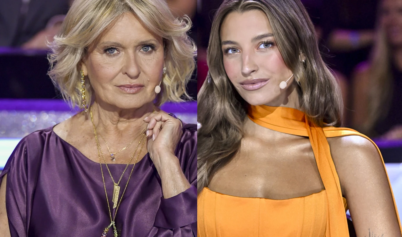 Ewa Kasprzyk i Julia Wieniawa Dwie kobiety siedzące obok siebie, jedna ubrana w fioletową sukienkę z kilkoma złotymi naszyjnikami, druga w pomarańczową sukienkę z odsłoniętymi ramionami, obie z założonymi mikrofonami na policzkach.