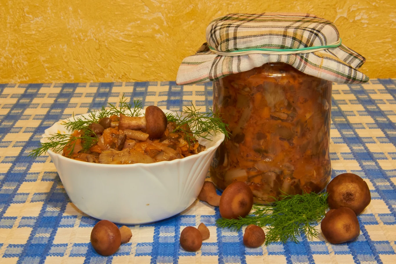 Grzyby po cygańsku robię jesienią na kilogramy. Smakują obłędnie Biała miska z duszonymi grzybami i koperkiem obok słoika z marynowanymi grzybami na niebiesko-białym obrusie.