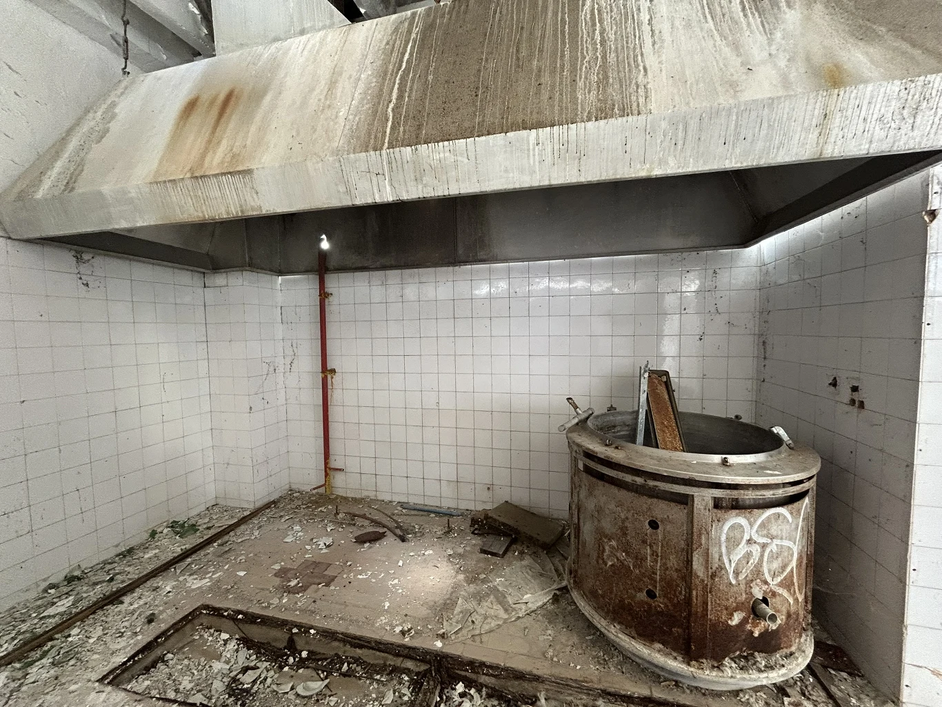 Kuchnia nieczynnego już ośrodka wczasowego/ Fot. Katarzyna Pruszkowska Stara, zdewastowana kuchnia przemysłowa z dużym, brudnym okapem, odsłoniętym czerwonym przewodem i zardzewiałym piecem, wokół którego leżą resztki gruzu.