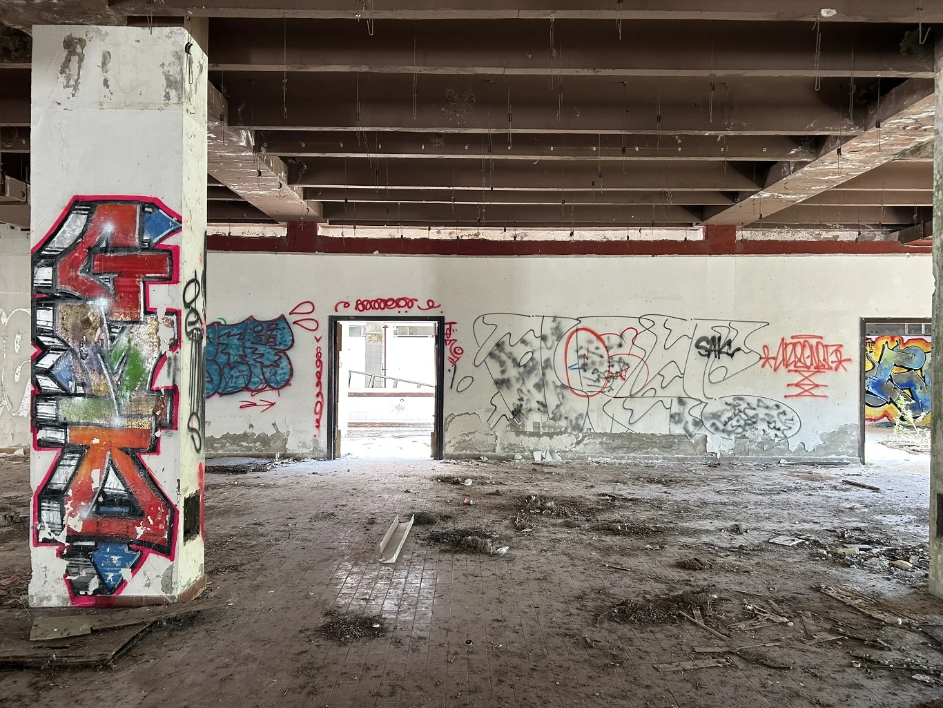 Ruiny dawnego ośrodka wczasowego/ Fot. Katarzyna Pruszkowska Opuszczone pomieszczenie z betonowym filarem i ścianami pokrytymi kolorowymi graffiti, z otwartymi drzwiami prowadzącymi na zewnątrz.