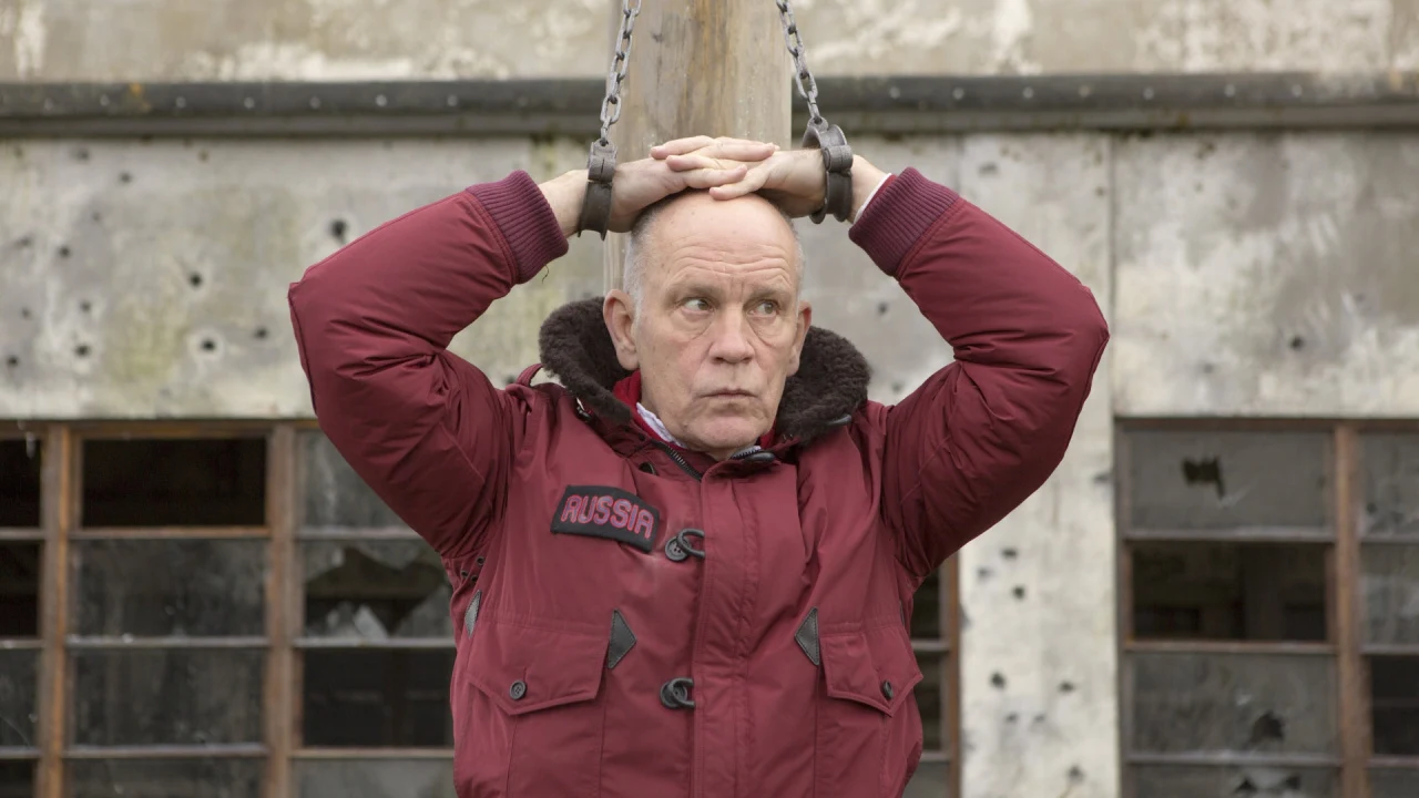 John Malkovich w filmie "Dziki Koń Dziewięć" DZIKI KOŃ DZIEWIĘĆ zr. Venice Film Festival Mężczyzna w bordowej kurtce z napisem 'Russia' stoi z rękami skrzyżowanymi nad głową, przywiązany do drewnianego słupa na tle zniszczonego budynku z wybitymi oknami.