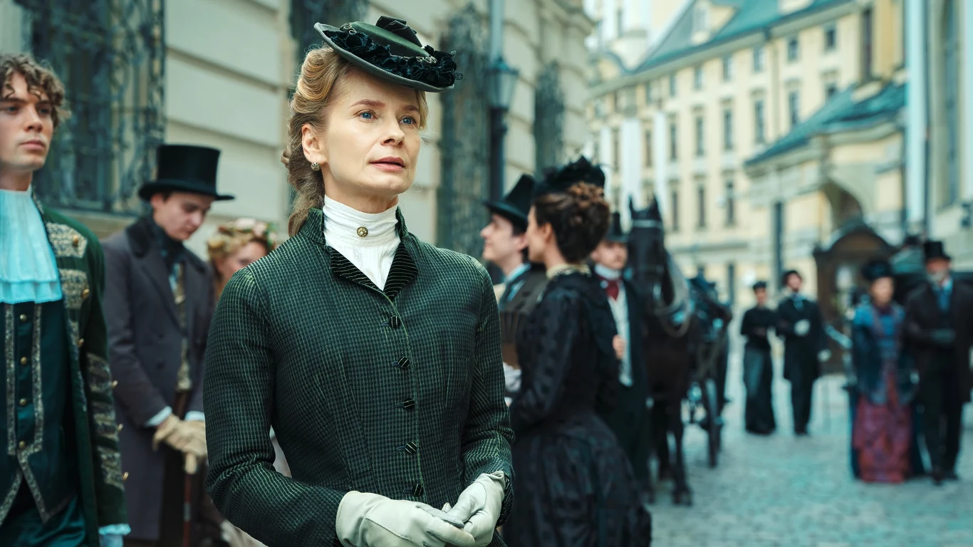 Magdalena Cielecka w roli Kazimiery Wąsowskiej Elegancko ubrana kobieta w stroju z epoki stoi na brukowanej ulicy w otoczeniu ludzi w historycznych kostiumach, na drugim planie widoczna zabytkowa architektura i dorożka z końmi.