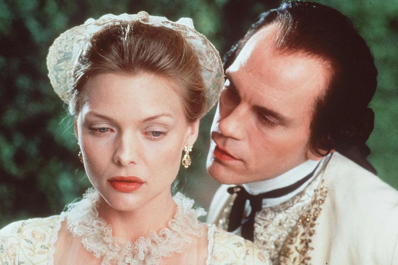 Z Michelle Pfeiffer w filmie "Niebezpieczne związki" Kobieta w białej sukni z koronką i perłowymi ozdobami odwraca wzrok z powagą, podczas gdy mężczyzna w eleganckim stroju z epoki pochyla się do jej ucha, sprawiając wrażenie wypowiadania sekretu.