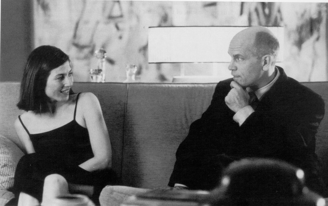 Kadr z filmu "Być jak John Malkovich" B Kobieta w ciemnej sukience i mężczyzna w garniturze siedzą obok siebie na kanapie, patrząc na siebie w skupieniu, z lampą stojącą za nimi.
