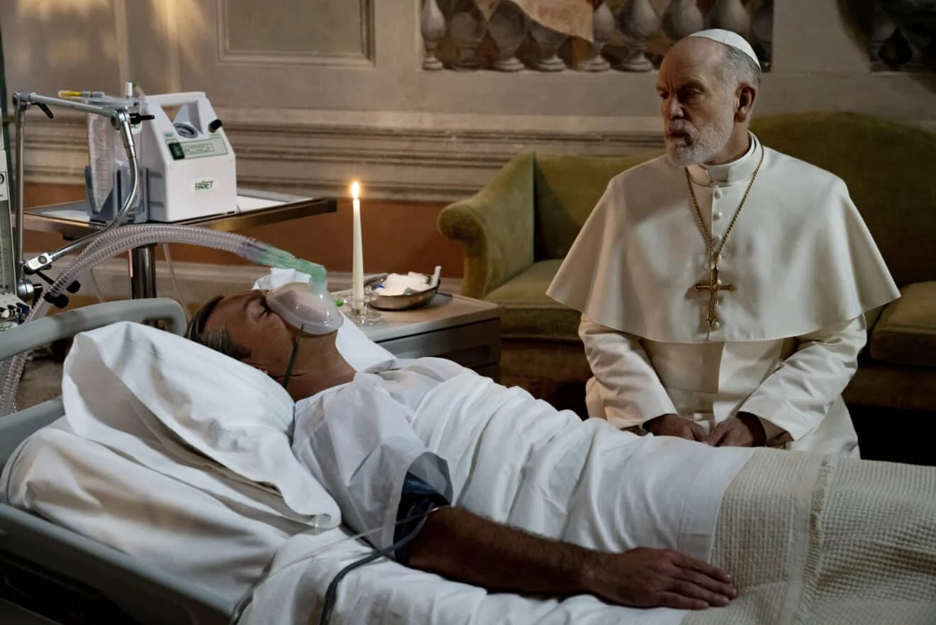 W serialu "Nowy papież" Mężczyzna w szpitalnym łóżku podłączony do respiratora, obok niego starszy mężczyzna w białych szatach i papieskiej piusce siedzący na krześle, w tle świeca i szpitalny sprzęt medyczny.