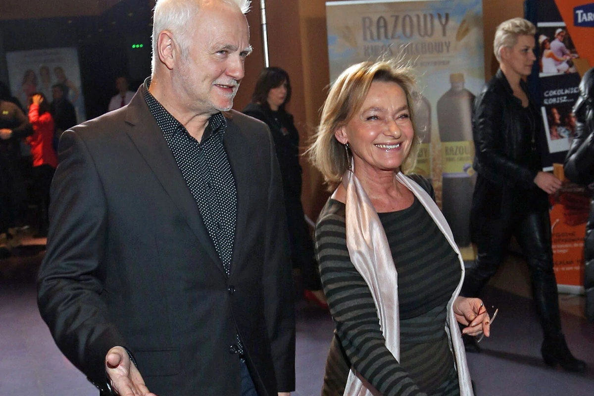 Krzysztof Piasecki i Anna Tomaszewska Uśmiechnięta para idąca razem, mężczyzna w ciemnej marynarce i kobieta w pasiastym swetrze z jasnym szalem, tło z reklamami i innymi osobami w półmroku.