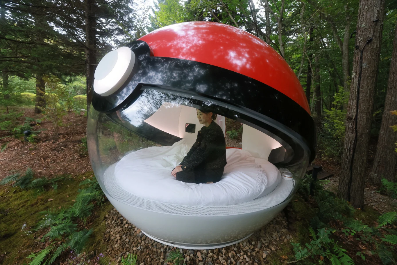 Hotel w kształcie Poke Balla Osoba siedząca na dużym, okrągłym łóżku umieszczonym w sferycznej kapsule przypominającej pokeballa, otoczonej zielonym lasem.