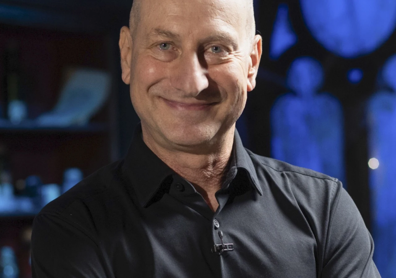 Wojciech Jagielski Wojciech Jagielski w czarnej koszuli, uśmiechnięty, patrzący w obiektyw na tle niebieskiego światła.