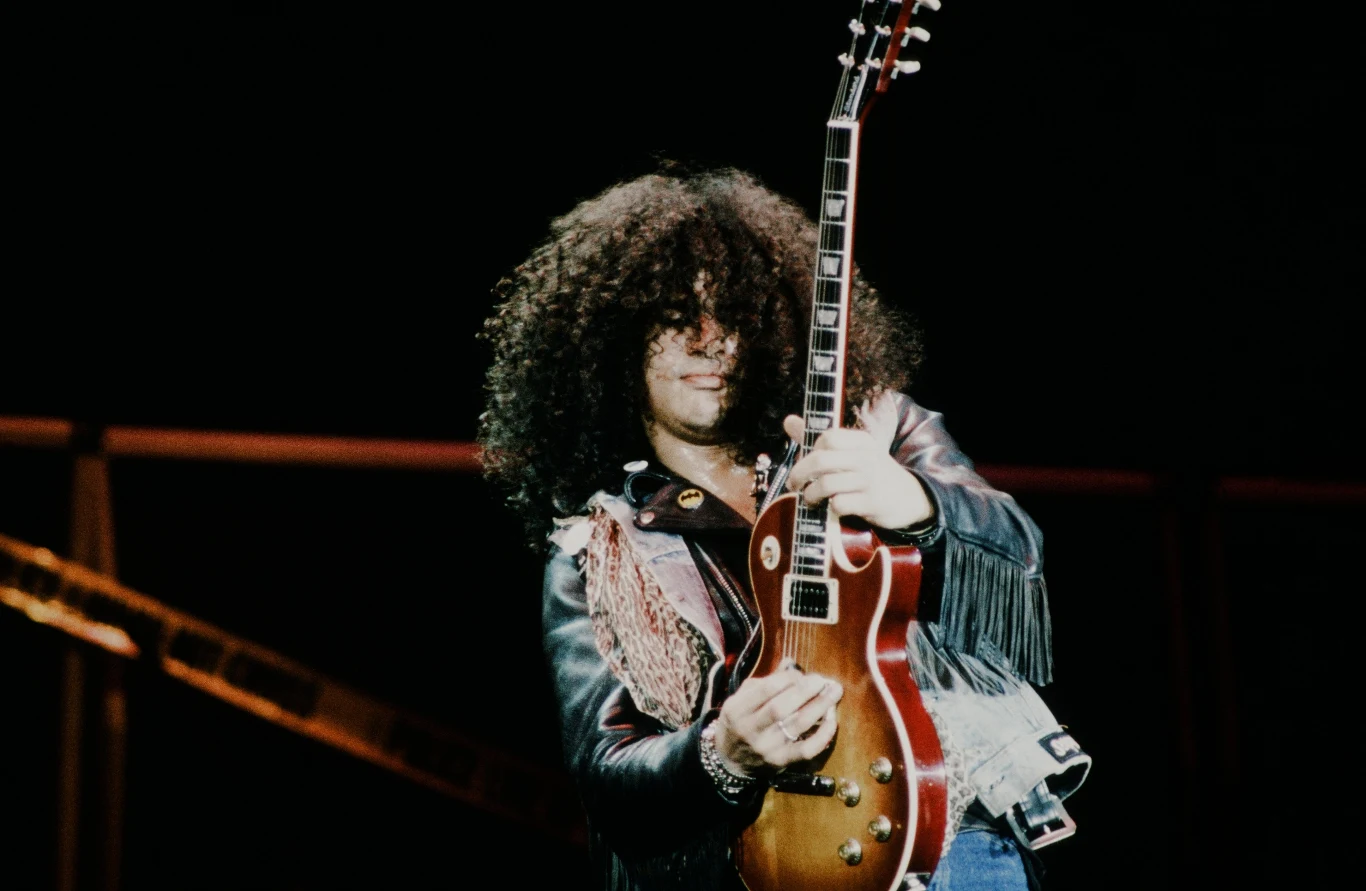Slash (Guns N' Roses) w 1991 r. Mężczyzna z bujnymi, kręconymi włosami grający na gitarze elektrycznej w skórzanej kurtce z frędzlami, skupiony na grze na scenie koncertowej.