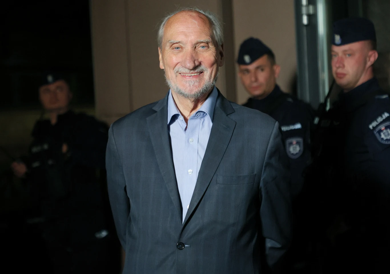 Poseł PiS Antoni Macierewicz przed Akademią Wymiaru Sprawiedliwości Antoni Macierewicz w garniturze przed Akademią Wymiaru Sprawiedliwości, w tle policjanci w mundurach.
