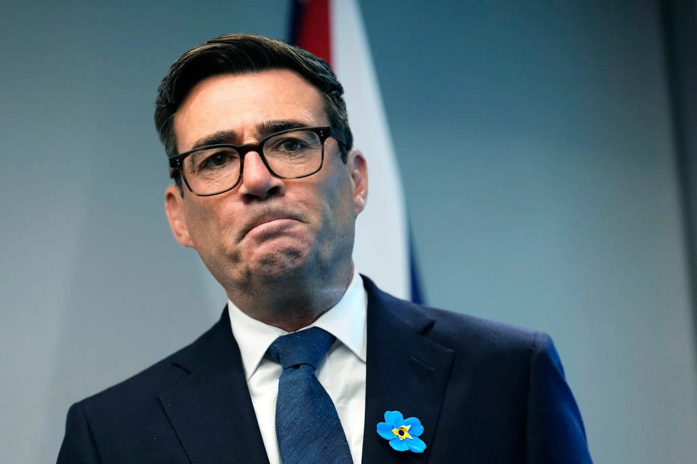 Andy Burnham, premier Wielkiej Brytanii Mężczyzna w okularach ubrany w ciemny garnitur, białą koszulę i niebieski krawat, z przypiętym niebieskim kwiatem do klapy; w tle fragment czerwono-biało-niebieskiej flagi.
