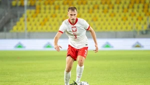 Piłkarz w białej koszulce z orłem i czerwonych spodenkach kontroluje piłkę na boisku podczas meczu na stadionie z żółtymi siedziskami.