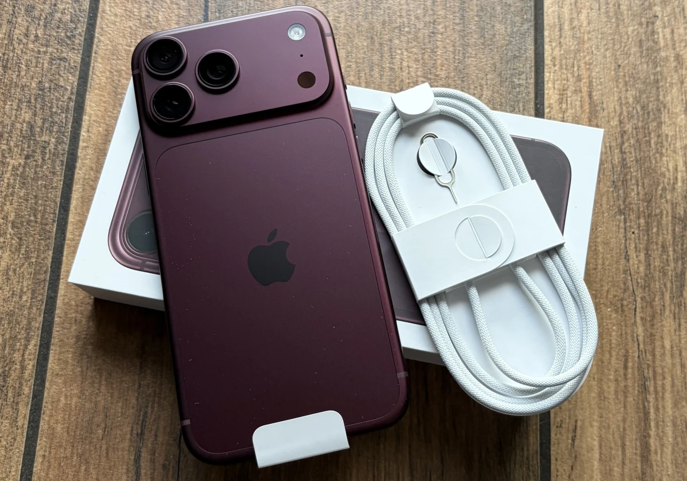 Zawartość opakowania ze smartfonem iPhone 18 Pro Max. Smartfon marki Apple w bordowej obudowie wraz z białym kablem do ładowania, narzędziem do wyjmowania tacki na kartę SIM oraz oryginalnym pudełkiem, wszystko umieszczone na drewnianej powierzchni.