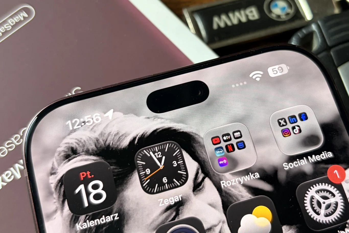 iPhone 18 Pro Max ma mniejszą dynamiczną wyspę. Górna część ekranu smartfona z widocznym zegarem, datą oraz ikonami aplikacji, w tle klucz samochodowy BMW i fragment etui telefonu.