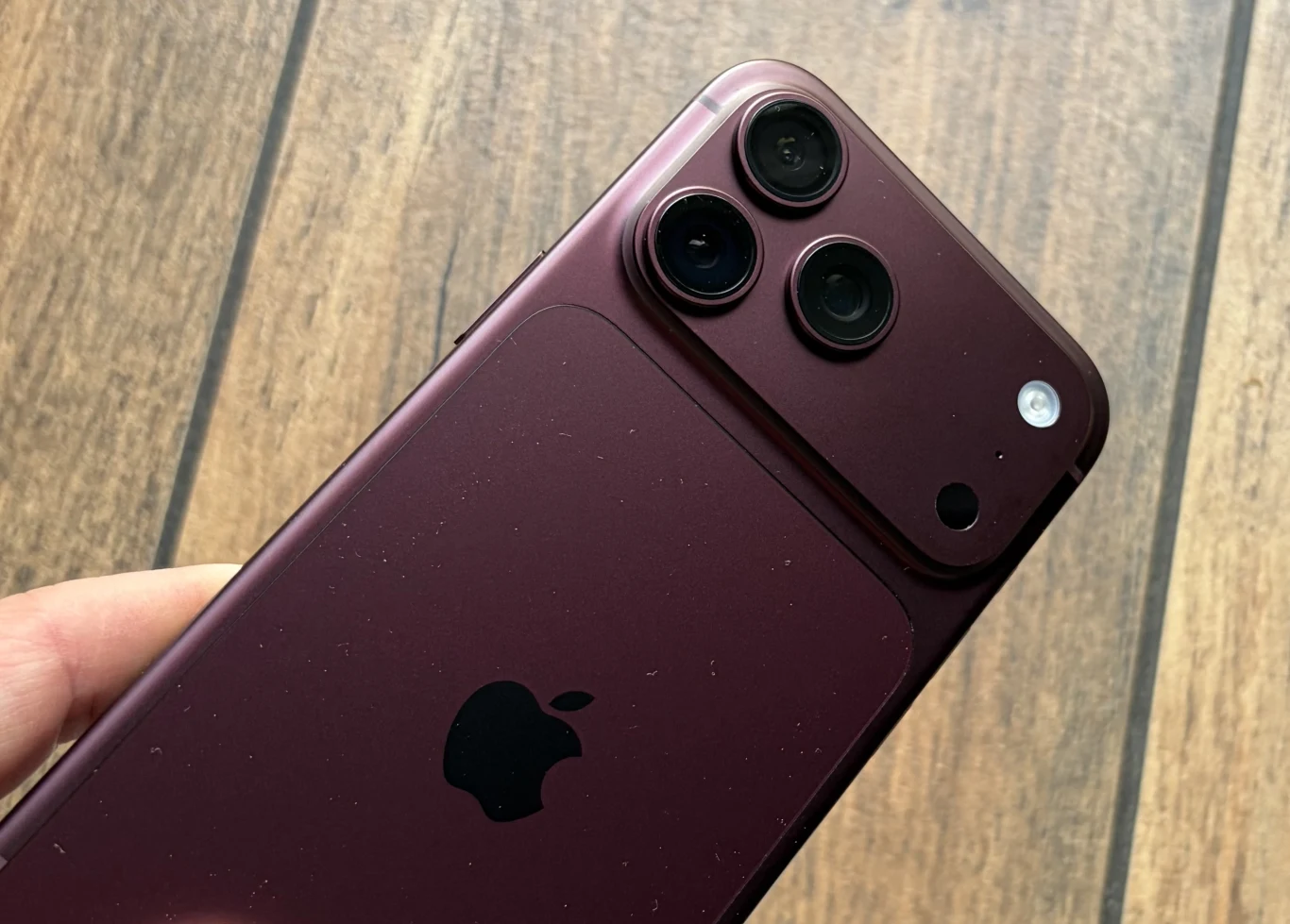 iPhone 18 Pro Max w kolorze burgundu. Bordowy smartfon marki Apple z trzema obiektywami aparatu i lampą błyskową, trzymany w dłoni na tle drewnianej podłogi.