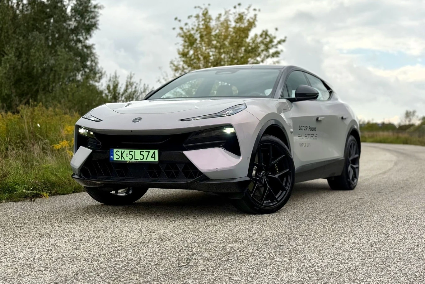 Lotus Eletre określany jest przez producenta jako "Hiper-SUV". Już wiem, dlaczego. Jasnoszary Lotus Eletre, sportowy SUV z czarnymi elementami, na zakręcie asfaltowej drogi wśród zieleni.