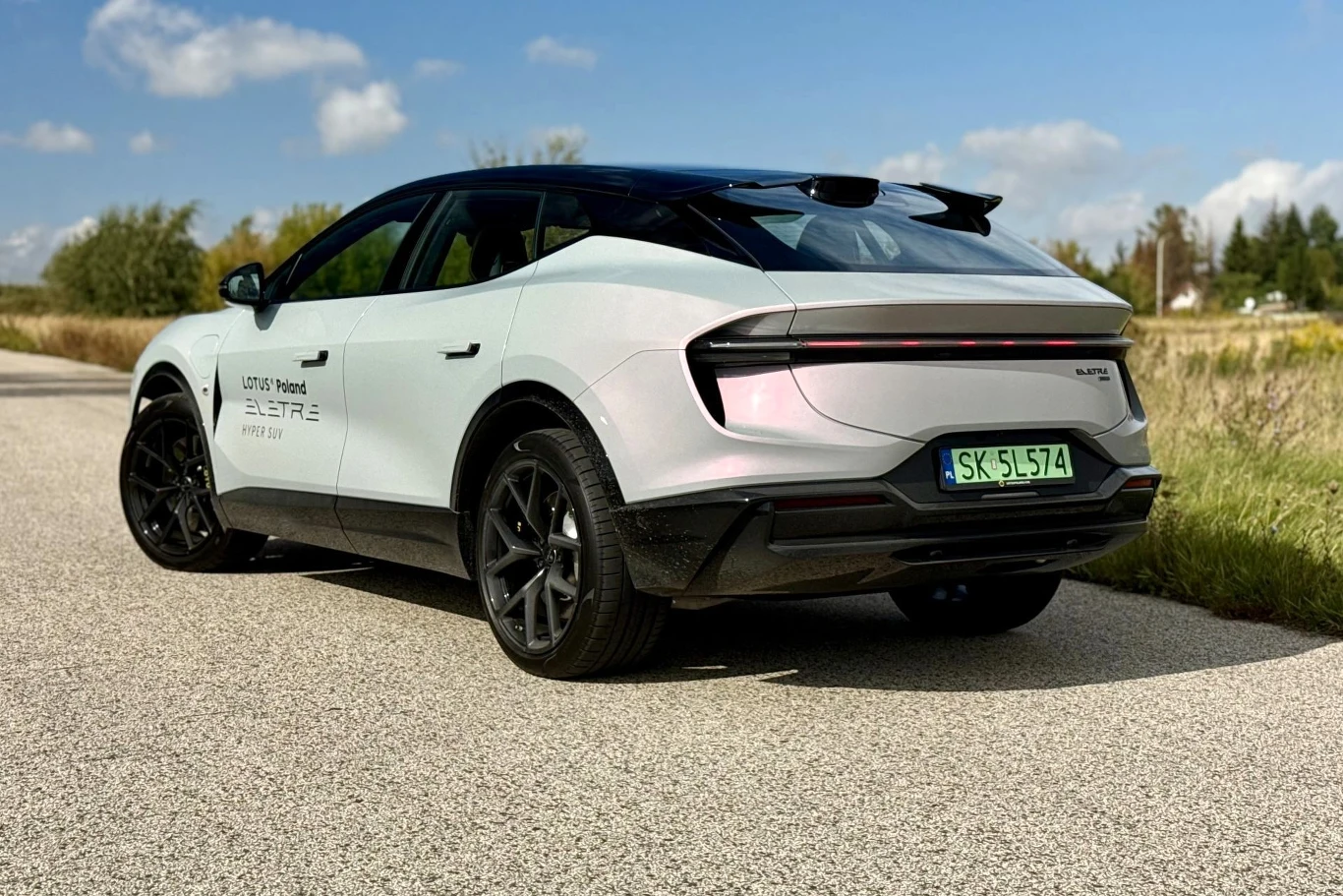 Tył Lotusa Eletre wygląda futurystycznie, jak gdyby był to model koncepcyjny. Biały elektryczny SUV Lotus Eletre z czarnym dachem i przyciemnianymi szybami stoi zaparkowany na wiejskiej drodze asfaltowej, prezentując nowoczesny, dynamiczny design oraz charakterystyczne tylne światła LED.