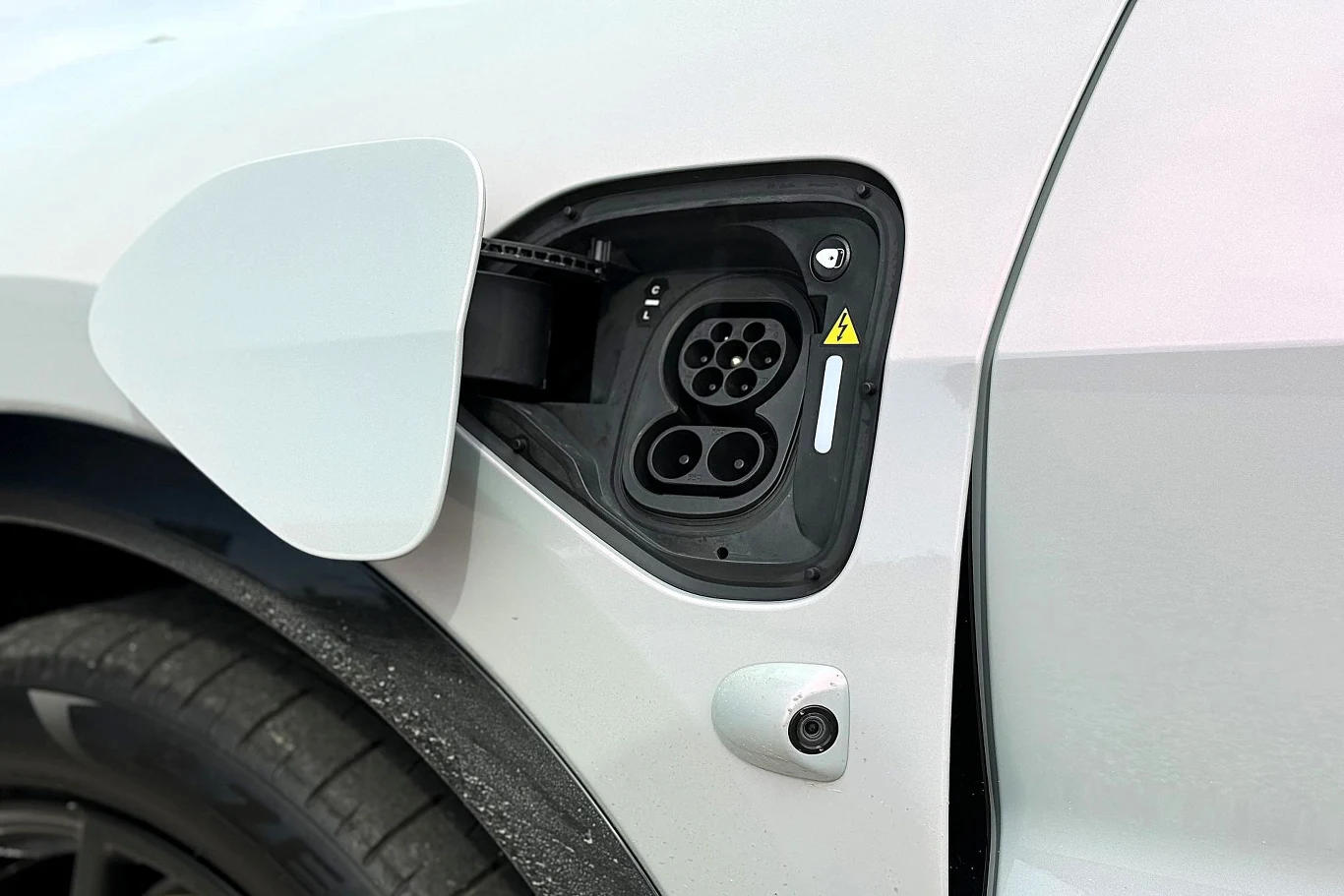 Akumulator o pojemności 107 kWh netto pozwala przejechać do 600 km na jednym ładowaniu. Zbliżenie na port ładowania samochodu elektrycznego z otwartą klapką, widoczne gniazdo do podłączenia kabla oraz tylna część opony pojazdu.
