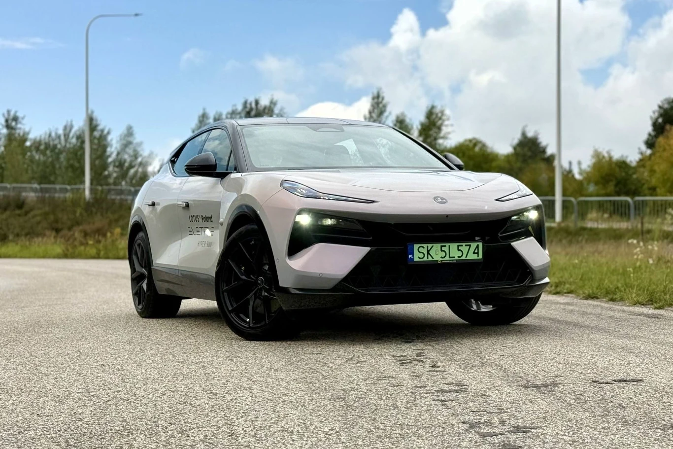 Lotus Eletre dostępny jest w dwóch wariantach mocy - 603 i 905 KM. W obu napęd trafia na cztery koła. Biało-czarny nowoczesny samochód elektryczny z charakterystycznymi reflektorami LED i polską tablicą rejestracyjną, zaparkowany na asfaltowej drodze na tle zielonych drzew.
