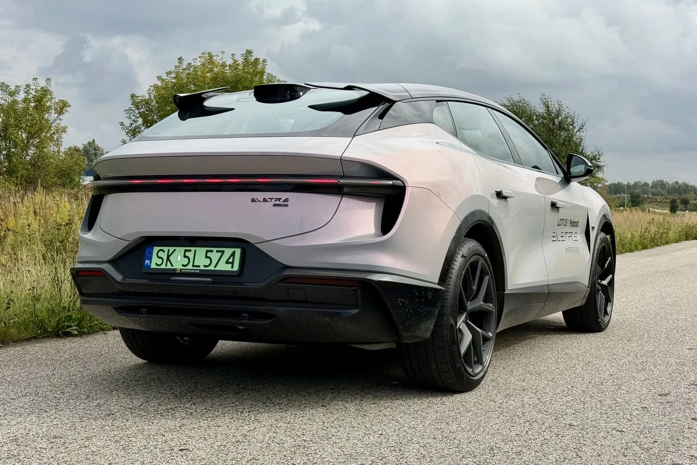 Lotus Eletre w wariancie 600 momentalnie reaguje na gaz, nawet w trybie zorientowanym na jak najdłuższy zasięg. Nowoczesny samochód elektryczny marki Lotus w kolorze srebrnym z czarnymi akcentami, widoczny z tyłu na pustej drodze, z charakterystycznym LED-owym pasem świetlnym oraz dużymi sportowymi felgami.
