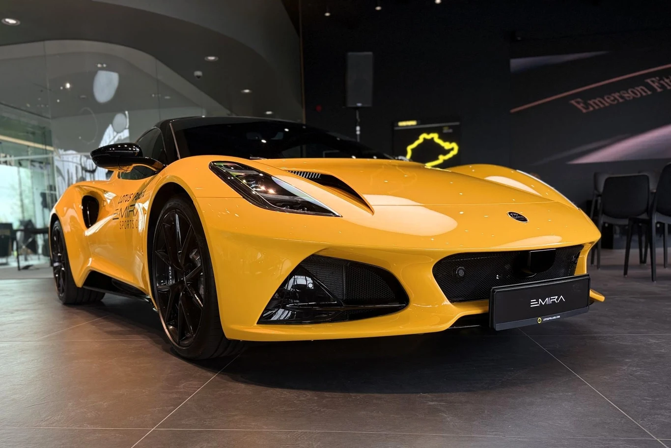 Lotus Emira dostępny jest z dwulitrowym czterocylindrowym silnikiem, ale też 3,5-litrowym V6. Żółty sportowy samochód Lotus Emira na czarnych felgach ustawiony w salonie samochodowym o nowoczesnym wystroju.