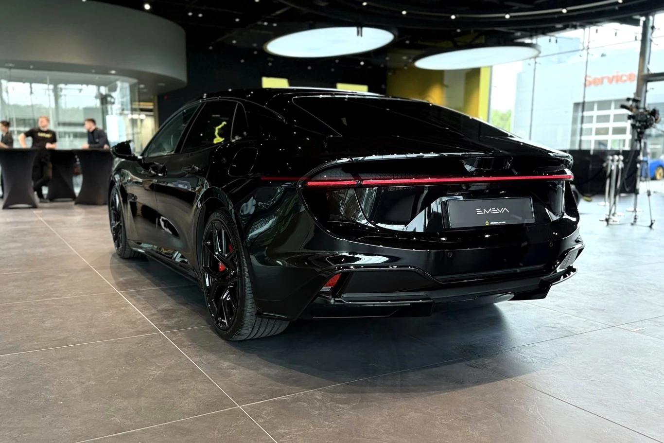 Lotus Emeya generuje do 918 KM. Czarny, nowoczesny sedan elektryczny o aerodynamicznej sylwetce, z wąską, ciągłą linią tylnych świateł oraz przyciemnianymi szybami, prezentowany w eleganckim salonie samochodowym.
