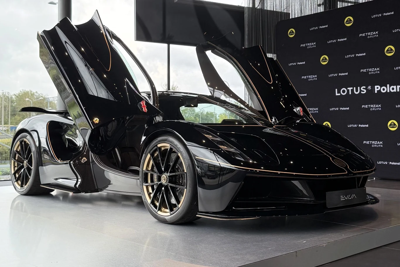 Lotus Evija wyposażony jest w cztery silniki elektryczne generujące łącznie 2039 KM. Czarny samochód sportowy Lotus z unoszonymi do góry drzwiami, złote felgi, wyeksponowany na tle ściany z logo marki.