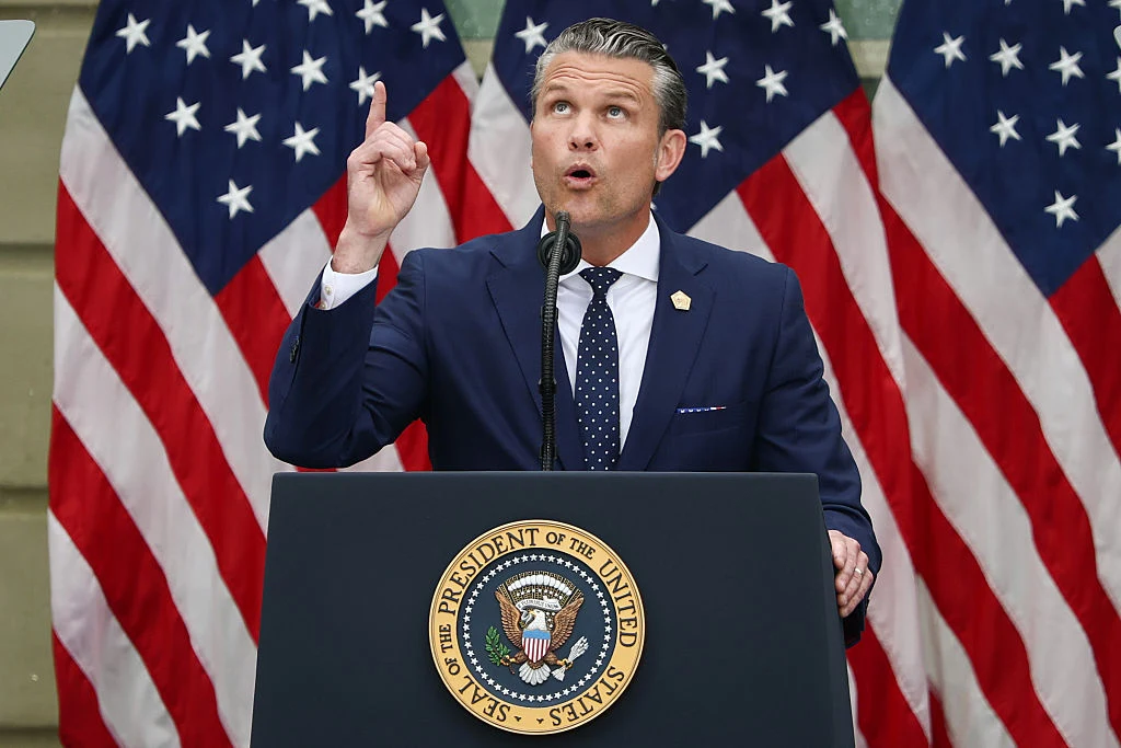Amerykański sekretarz wojny Pete Hegseth Pete Hegseth w granatowym garniturze przemawia przy mównicy z herbem prezydenta USA, w tle flagi amerykańskie