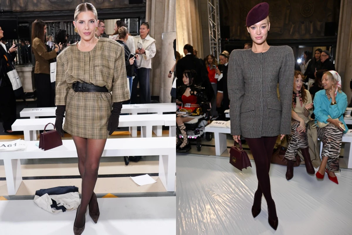 Dwie kobiety w stylowych, kraciastych marynarkach i ciemnych rajstopach, z małymi torebkami w dłoni, pozują na tle białych ławek w eleganckim wnętrzu podczas pokazu mody.
