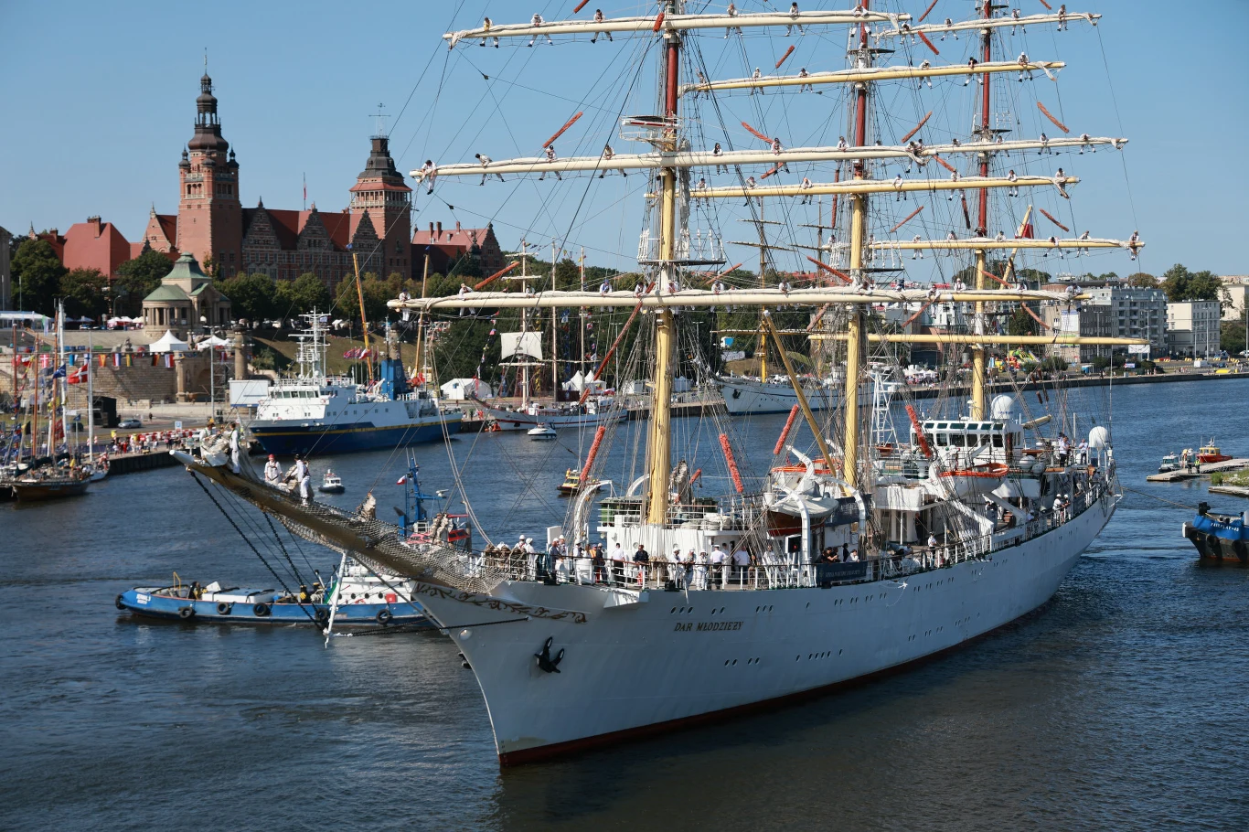Finał regat The Tall Ship Racec w 2024 roku. Dar Młodzieży w Szczecinie biały żaglowiec z wieloma masztami płynie po rzece, w tle okazały ceglany budynek z wieżami oraz liczne mniejsze łodzie na wodzie