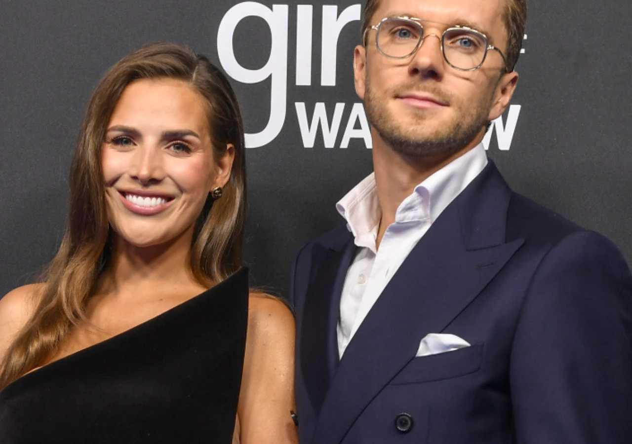 Ola Nowak, Maciej Mazurek Para ubrana elegancko, kobieta w czarnej sukni na jedno ramię, mężczyzna w granatowym garniturze i okularach, w tle ciemna ścianka z białymi napisami.