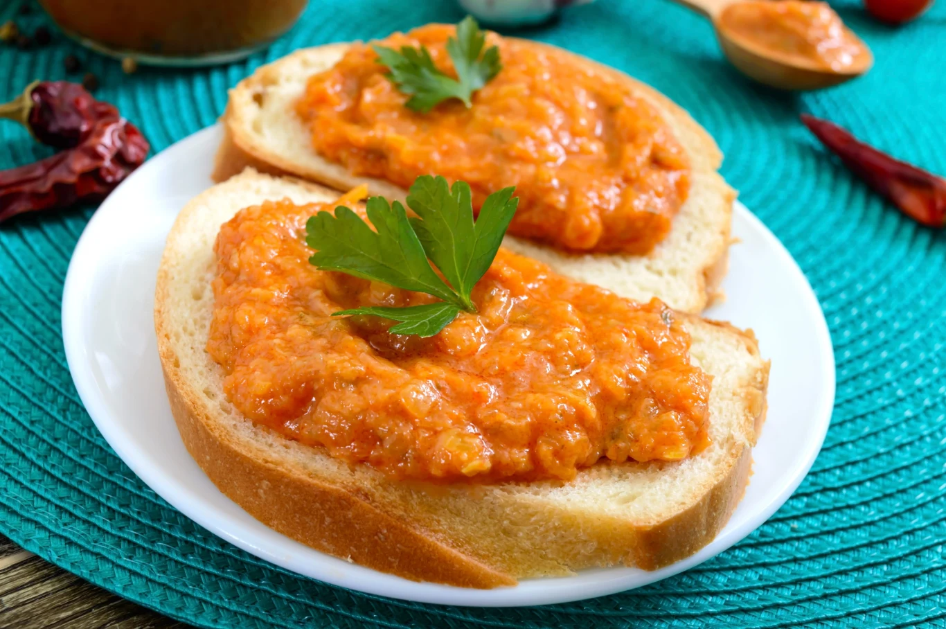 Ajwar ląduje u mnie na kanapkach, ale nie tylko Dwie kromki jasnego chleba z pastą ajvar i natką pietruszki na białym talerzu.