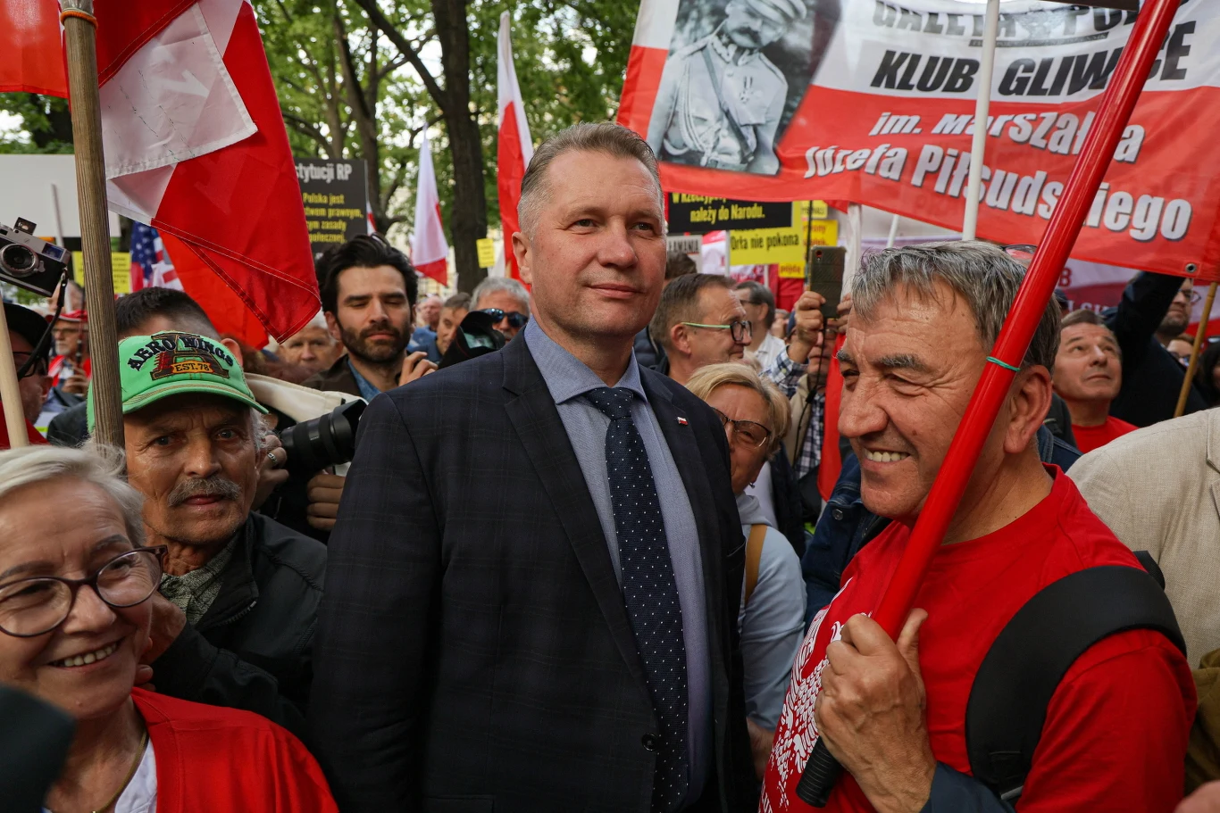 Uśmiechnięci ludzie zgromadzeni na wiecu z biało-czerwonymi flagami i transparentami oraz mężczyzna w garniturze stojący w centrum tłumu.