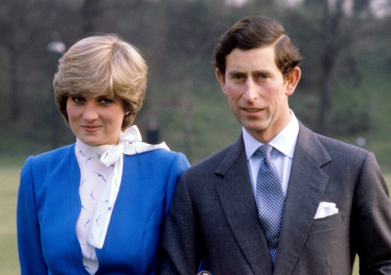 Księżna Diana i książę Karol w 1981 roku Kobieta w eleganckim niebieskim żakiecie z białą bluzką i mężczyzna w szarym garniturze z krawatem stoją obok siebie na świeżym powietrzu, patrząc w stronę aparatu.
