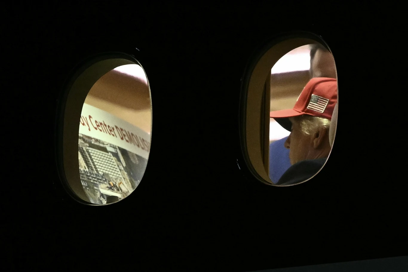 Donald Trump na pokładzie Air Force One z ilustracją, która prawdopodobnie zawiera napis "Kennedy Center DEMOLISHED" Mężczyzna w czerwonej czapce z naszywką w kształcie flagi USA widoczny przez okno samolotu, obok gazeta lub ulotka z napisem.