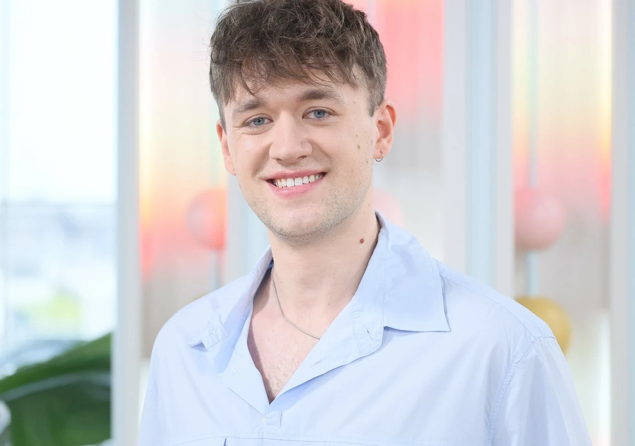 Mateusz Glen Młody mężczyzna z jasnymi oczami i krótkimi, brązowymi włosami uśmiecha się szeroko, ubrany w jasnoniebieską koszulę z rozpiętym kołnierzem, w tle jasne, rozmyte kolory.