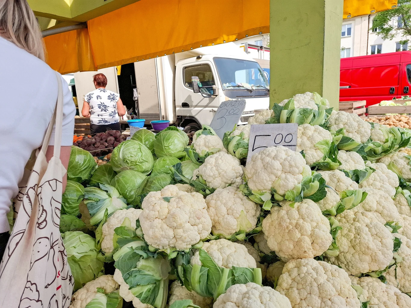 Kalafiory na łódzkim ryneczku Stos warzyw z przewagą świeżych kalafiorów i główek kapusty, pośrodku widoczna cena, w tle samochody i dwie osoby przy straganie.