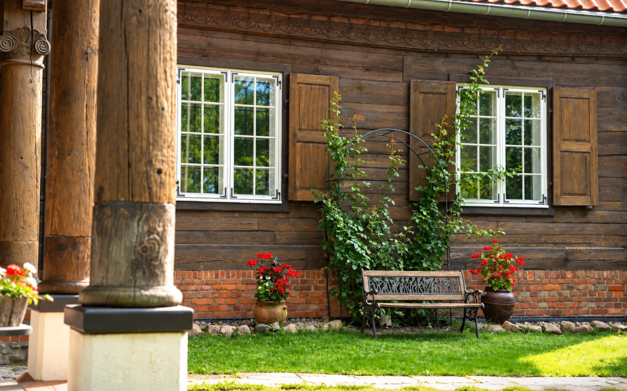 Dom w Marynowach Drewniana ławka ustawiona pod ścianą tradycyjnego domu z drewna, otoczona donicami z kwitnącymi czerwonymi pelargoniami.