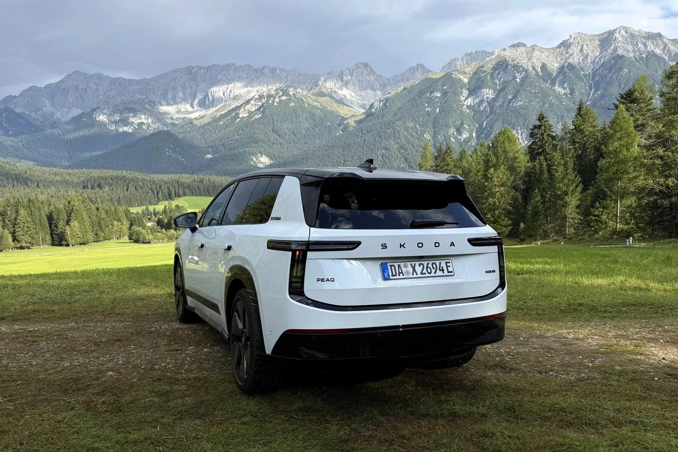 Skoda Peaq ma 4,87 m długości, o 11,5 cm więcej od Kodiaqa Biały SUV marki Skoda z czarnym dachem stoi na trawiastym terenie, w tle rozciągają się górskie szczyty i gęsty las.