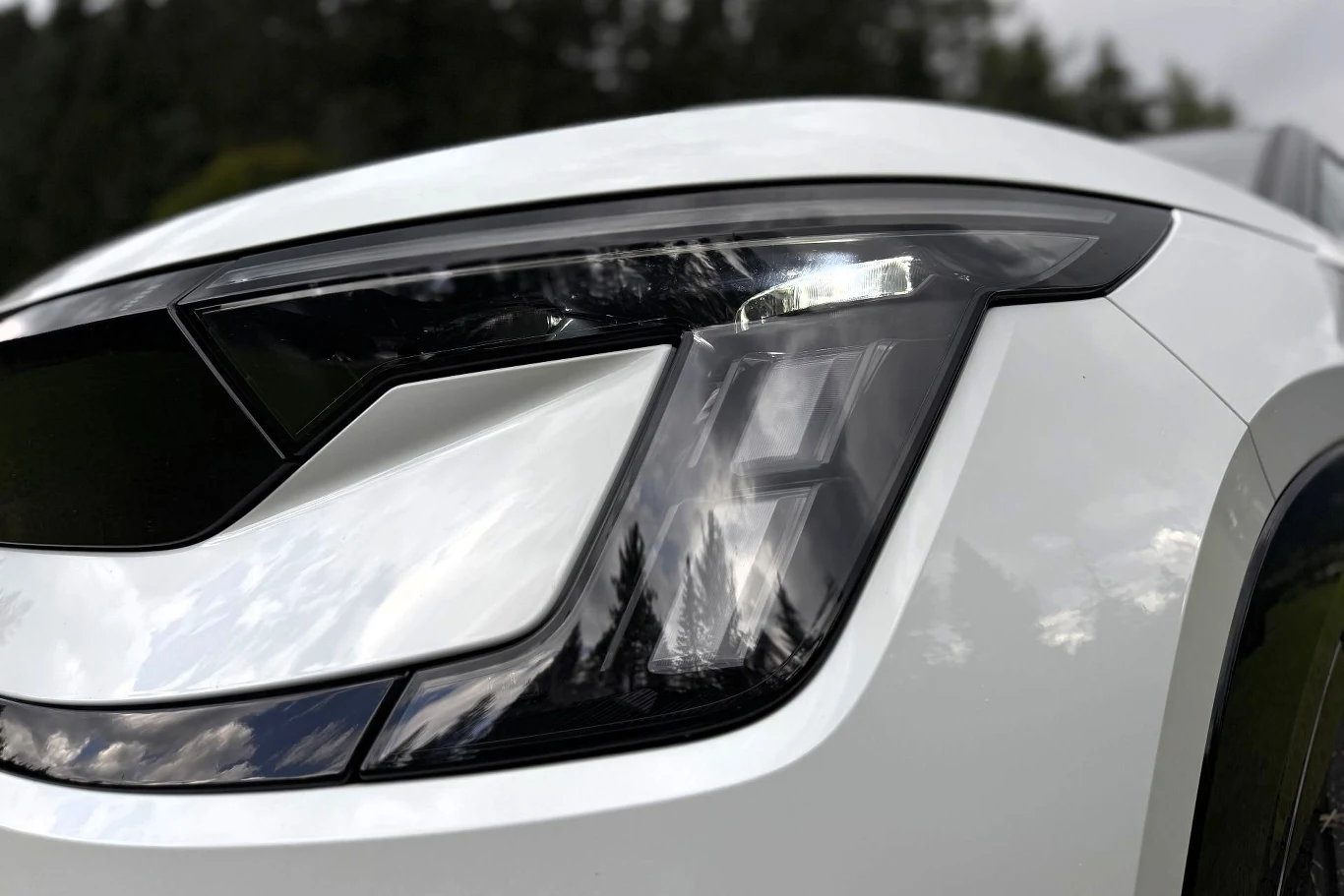 Skoda Peaq opcjonalnie może być wyposażona w reflektory Matrix LED. Nowoczesny reflektor LED w białym samochodzie, z wyraźnymi przetłoczeniami karoserii i odbiciem nieba na lakierze.