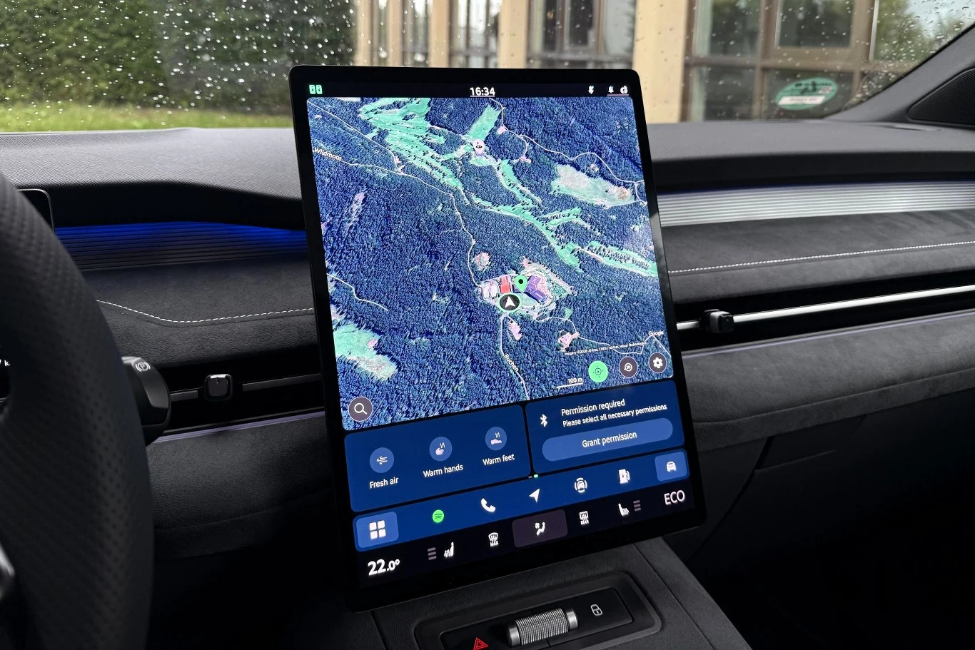 Nawigacja jest zintegrowana z bazą Google, co oznacza, że mamy dostęp do stale aktualizowanej listy miejsc z Map Google wraz z ich ocenami czy godzinami otwarcia. Duży, pionowy ekran dotykowy wyświetlający mapę satelitarną nawigacji samochodowej z widocznymi szczegółami leśnego terenu, umieszczony na nowoczesnej desce rozdzielczej auta.