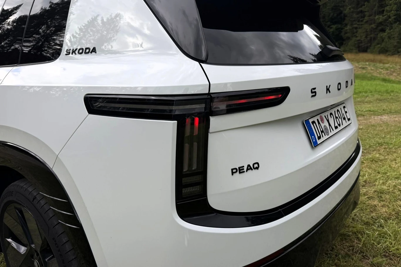 Skoda Peaq bardzo dobrze tłumi nierówności. Tył białego samochodu marki Škoda z widocznym logo, nowoczesnymi poziomymi lampami LED oraz oznaczeniem modelu PEAQ, ustawiony na trawie przy lesie.