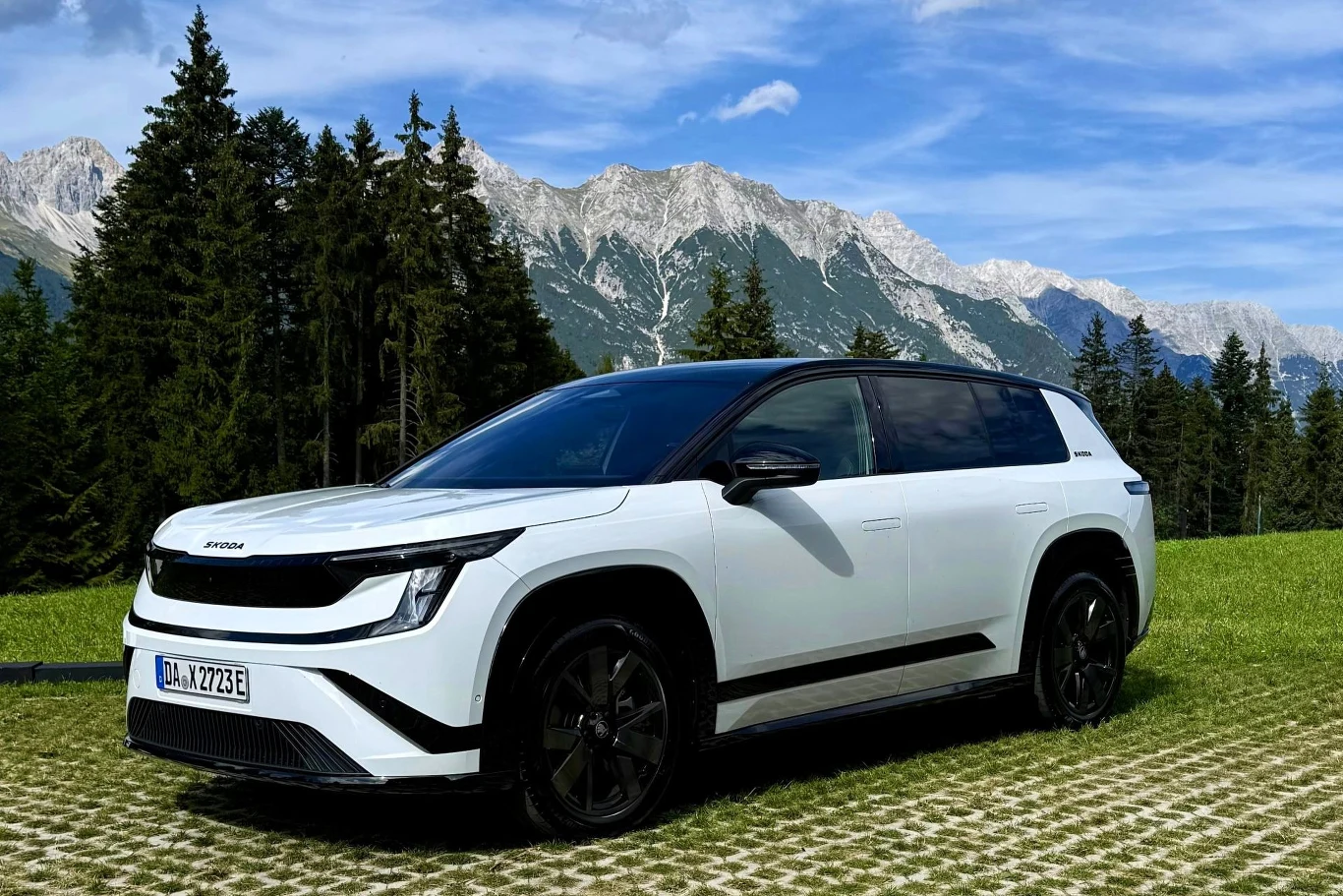 Skoda Peaq kosztuje minimum 204 900 zł. Biały samochód elektryczny SUV zaparkowany na zielonej trawie z czarnymi felgami, na tle lasu świerkowego i górskich szczytów pod błękitnym niebem.