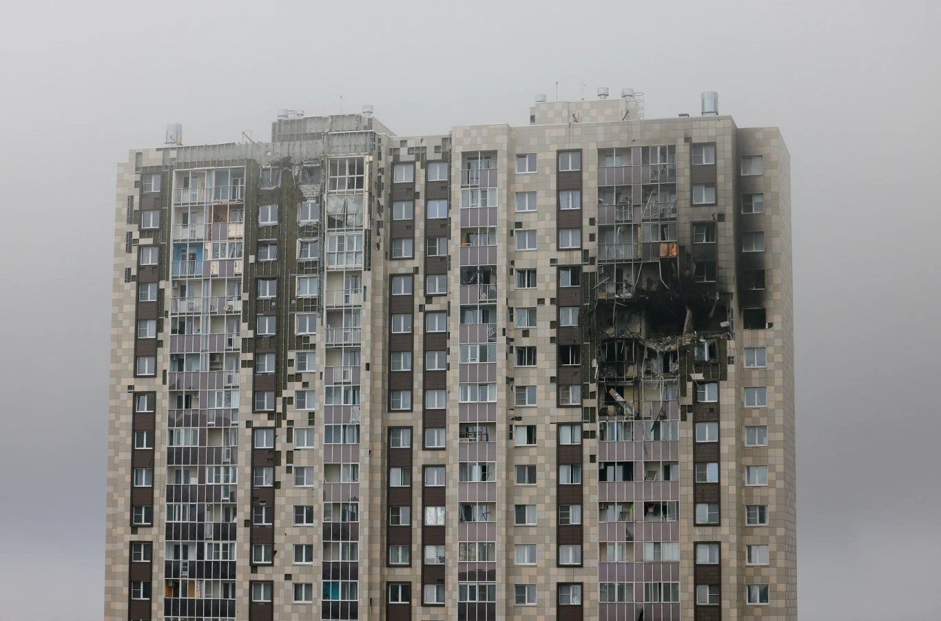 Wielopiętrowy blok mieszkalny z poważnie uszkodzonymi i osmalonymi fragmentami elewacji, oknami wybitymi na kilku piętrach oraz widocznymi zniszczeniami konstrukcji budynku.