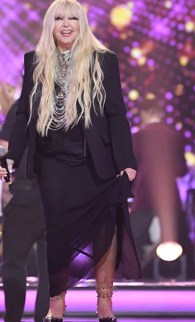 Maryla Rodowicz w "Tańcu z gwiazdami" Kobieta o długich blond włosach ubrana w czarną, elegancką suknię z żakietem oraz licznymi srebrnymi naszyjnikami, stojąca na scenie z mikrofonem w dłoni na tle kolorowych świateł.