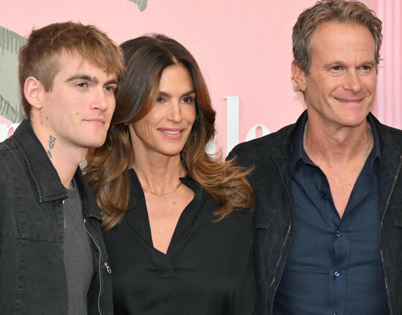 Presley Gerber wraz z rodzicami, Cindy Crawford i Rande Gerberem. Trzy uśmiechnięte osoby stoją obok siebie, ubrane w ciemne, casualowe ubrania, patrząc przed siebie na tle jasnoróżowej ściany.