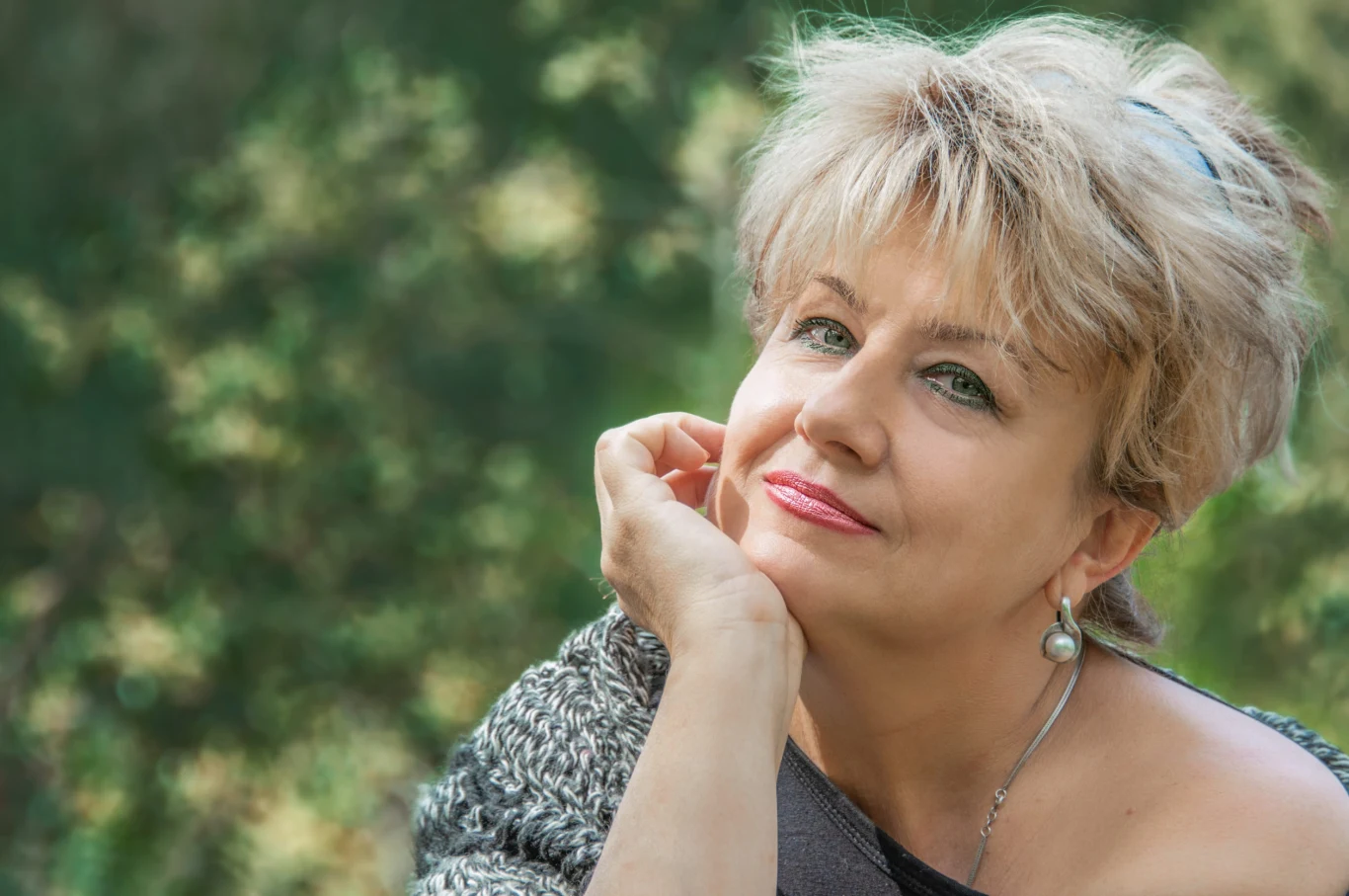 Uśmiechnięta kobieta w średnim wieku z krótkimi, jasnymi włosami opiera głowę na dłoni, nosząc srebrny kolczyk i sweter, patrzy w stronę aparatu na tle zieleni.