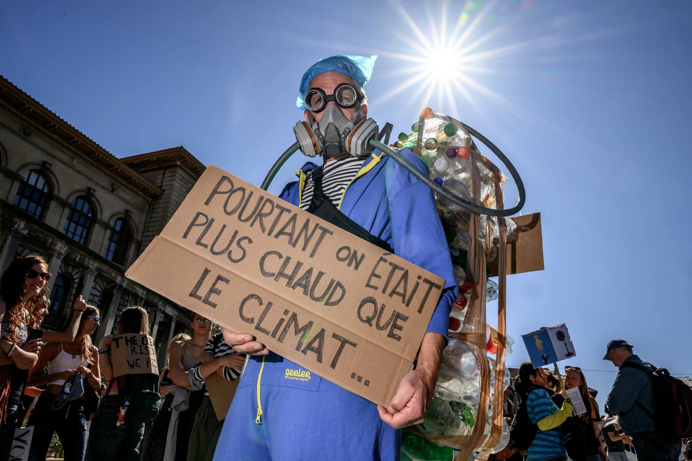 Protesty klimatyczne w Szwajcarii mają potrwać co najmniej do października Osoba w niebieskim stroju i masce gazowej trzyma tablicę z francuskim napisem, mając na plecach konstrukcję z butelek plastikowych, w otoczeniu protestujących na tle jasnego słońca.